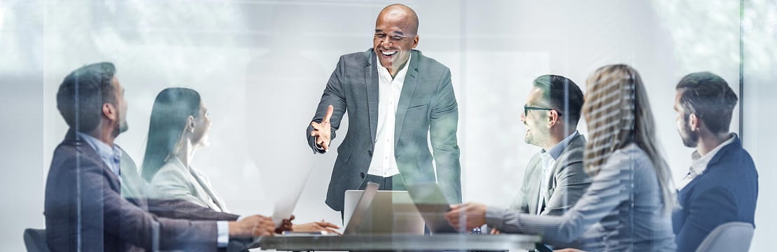 Man stands at the head of a table and is speaking with five individuals seated around the table from him